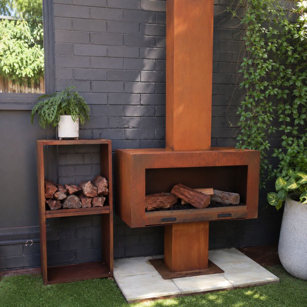 Corten Steel Grand Hearth Fireplace in a contemporary outdoor entertaining area, featuring a rust-finished steel wood-burning fireplace with a tall chimney and spacious firebox. Positioned beside a matching corten steel log storage unit against a modern brick wall, creating a warm and stylish backyard focal point.