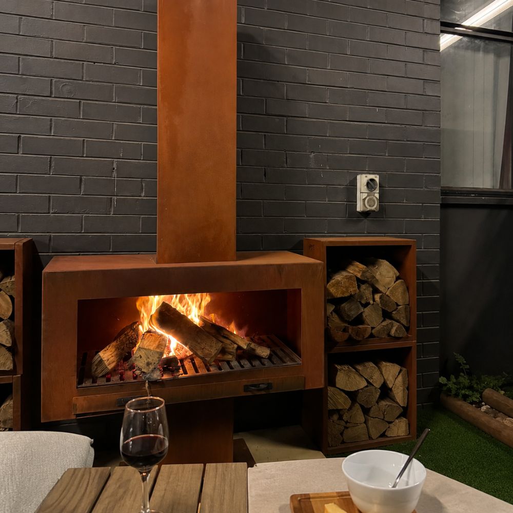 Corten Steel Grand Hearth Fireplace in a modern outdoor entertaining space, featuring a rust-finished steel wood-burning fireplace with a tall chimney and roaring fire. Framed by matching corten steel log storage units and set beside an outdoor dining area, it creates a warm, inviting atmosphere for relaxing and entertaining.