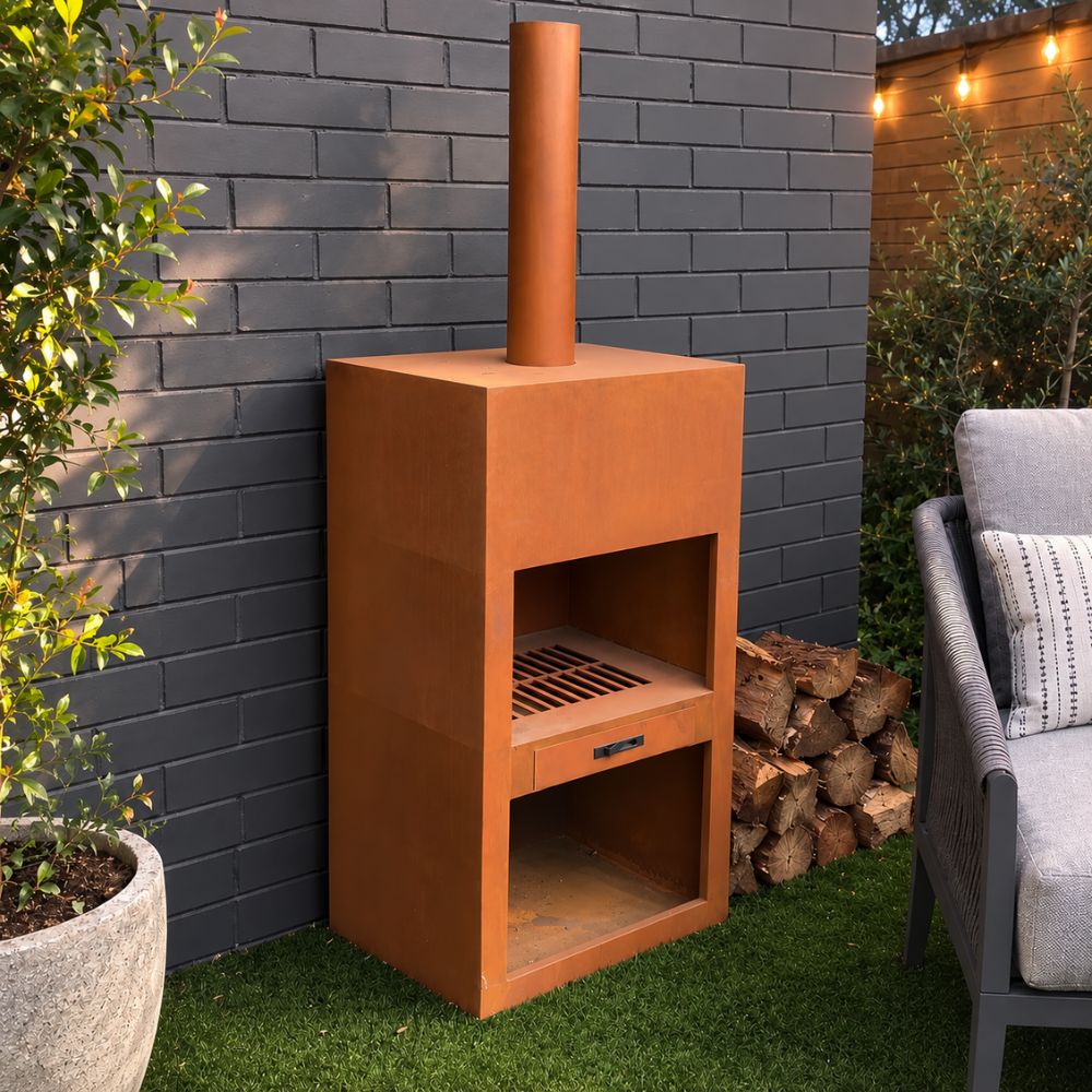 Corten Steel Heat Haven Fire Place in a modern outdoor entertaining area, featuring a rust-finished steel wood-burning fireplace with a tall chimney, spacious firebox, and integrated lower storage compartment. Positioned against a contemporary brick wall beside stacked firewood and outdoor seating, it offers a stylish focal point for year-round backyard entertaining.