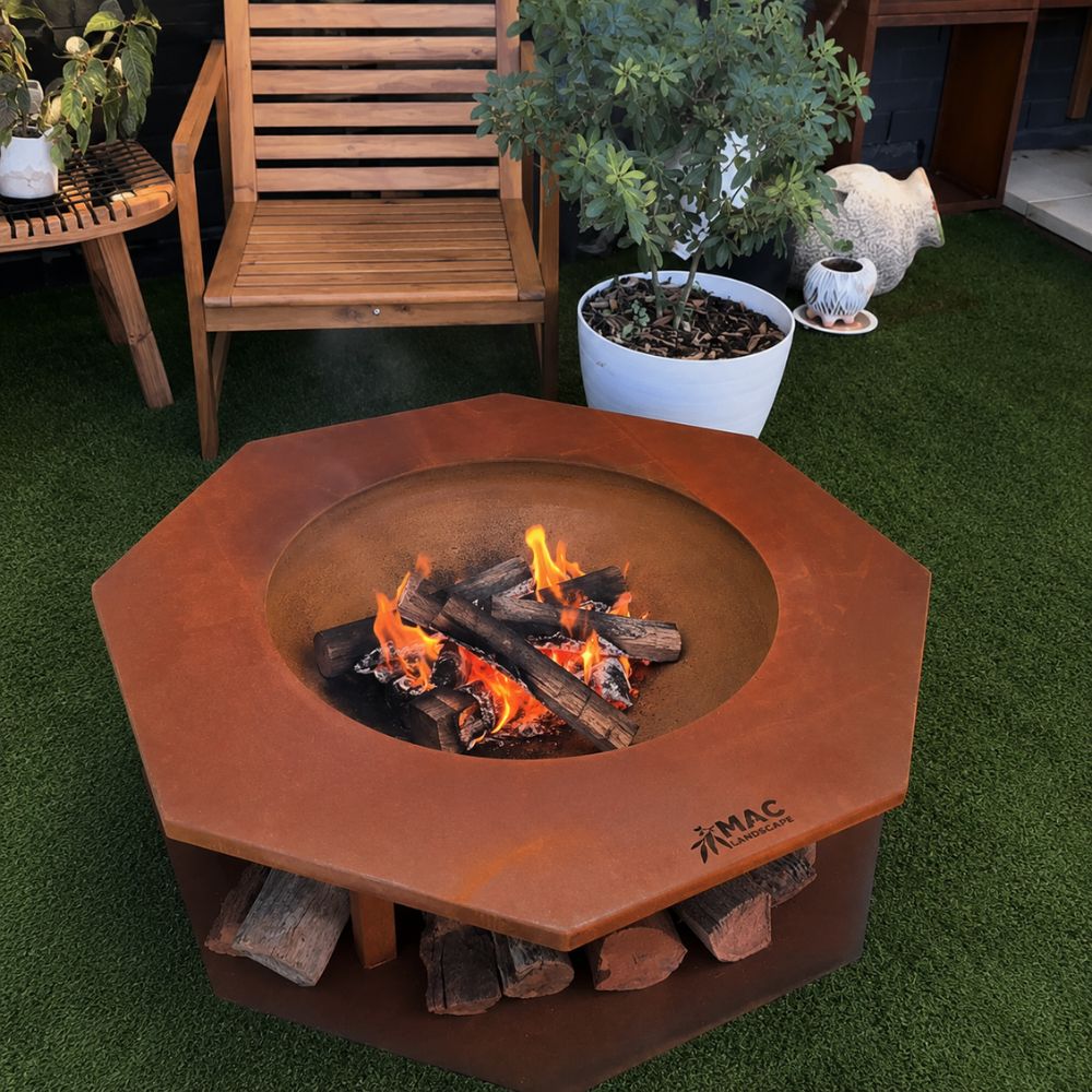 Corten Steel King Midus Fire Pit in a contemporary outdoor entertaining area, featuring a rust-finished octagonal steel fire pit with a wide tabletop surround and integrated firewood storage underneath. The wood-burning fire creates a warm, inviting focal point, complemented by timber outdoor furniture and surrounding garden planters.