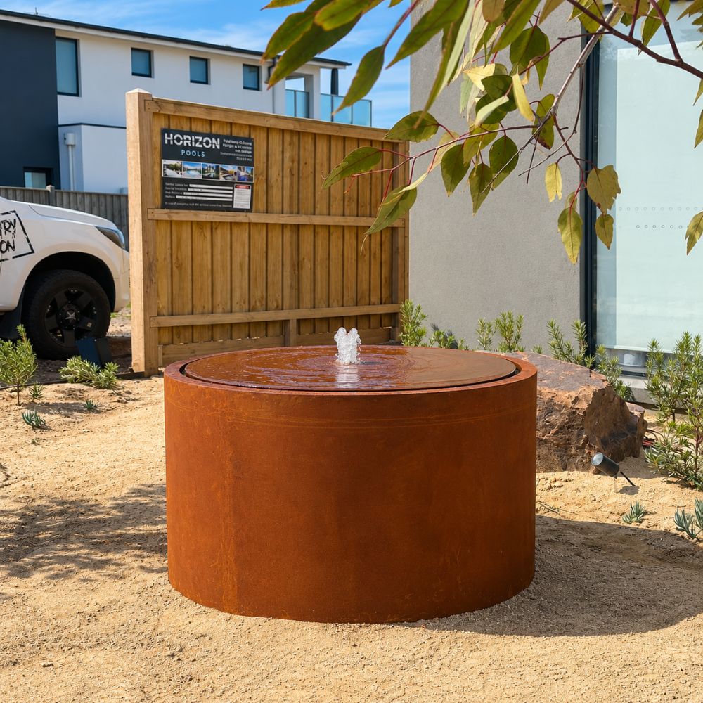Corten Steel Rita Round Narrow Water Fountain in a contemporary front garden, featuring a circular rust-finished steel fountain with a central bubbling water feature. Surrounded by low-maintenance landscaping, sandy garden beds, natural stone, and modern residential architecture.