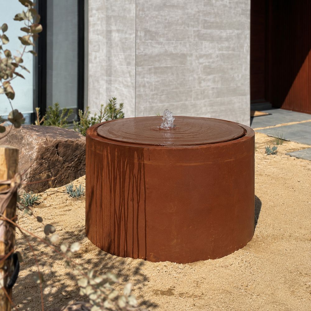 Corten Steel Rita Round Narrow Water Fountain in a contemporary landscape, featuring a cylindrical rust-finished steel fountain with a bubbling central water feature. Set within a sandy garden with natural boulders, drought-tolerant plants, and a modern home façade, creating a minimalist outdoor focal point.