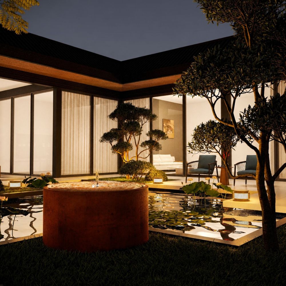 Corten Steel Rita Round Narrow Water Fountain with Light in a luxury outdoor courtyard at night, featuring a rust-finished cylindrical steel fountain with an illuminated bubbling water feature. Positioned beside a reflective pond with water lilies, landscaped trees, and a modern home, creating a warm and elegant garden ambience.