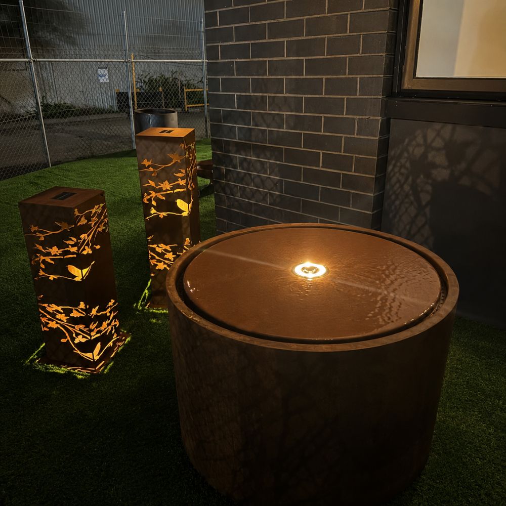 Corten Steel Rita Round Narrow Water Fountain with Light in a modern outdoor setting at night, featuring a circular rust-finished steel fountain with an illuminated central bubbling water feature. Surrounded by decorative corten steel garden lights, creating a warm ambient glow against contemporary landscaping and architecture.