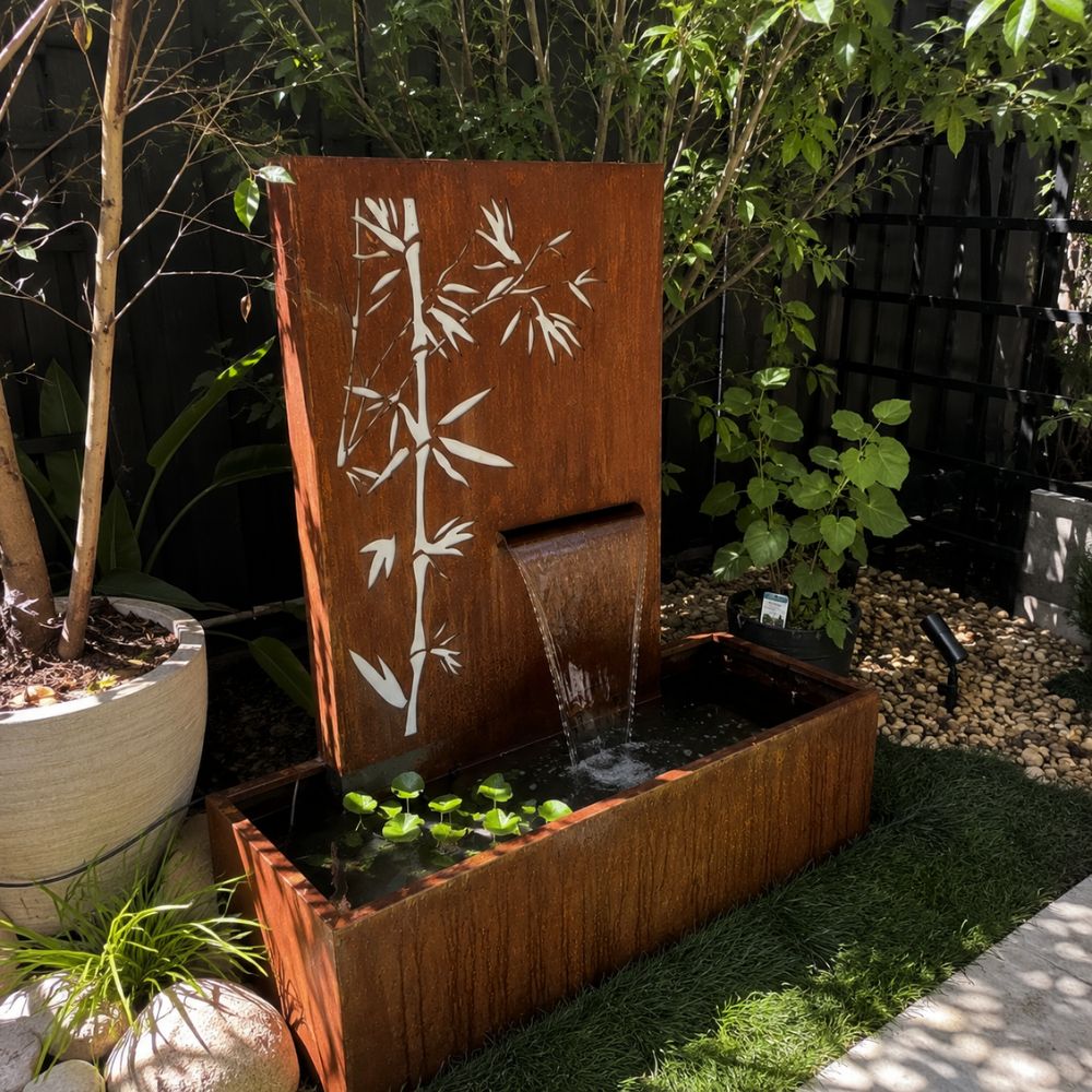 Corten Steel Serenity Water Fountain featuring a rust-finished steel waterfall with an elegant laser-cut bamboo design. Water cascades smoothly into a matching rectangular basin, creating a tranquil focal point in a contemporary garden surrounded by lush greenery and natural landscaping.