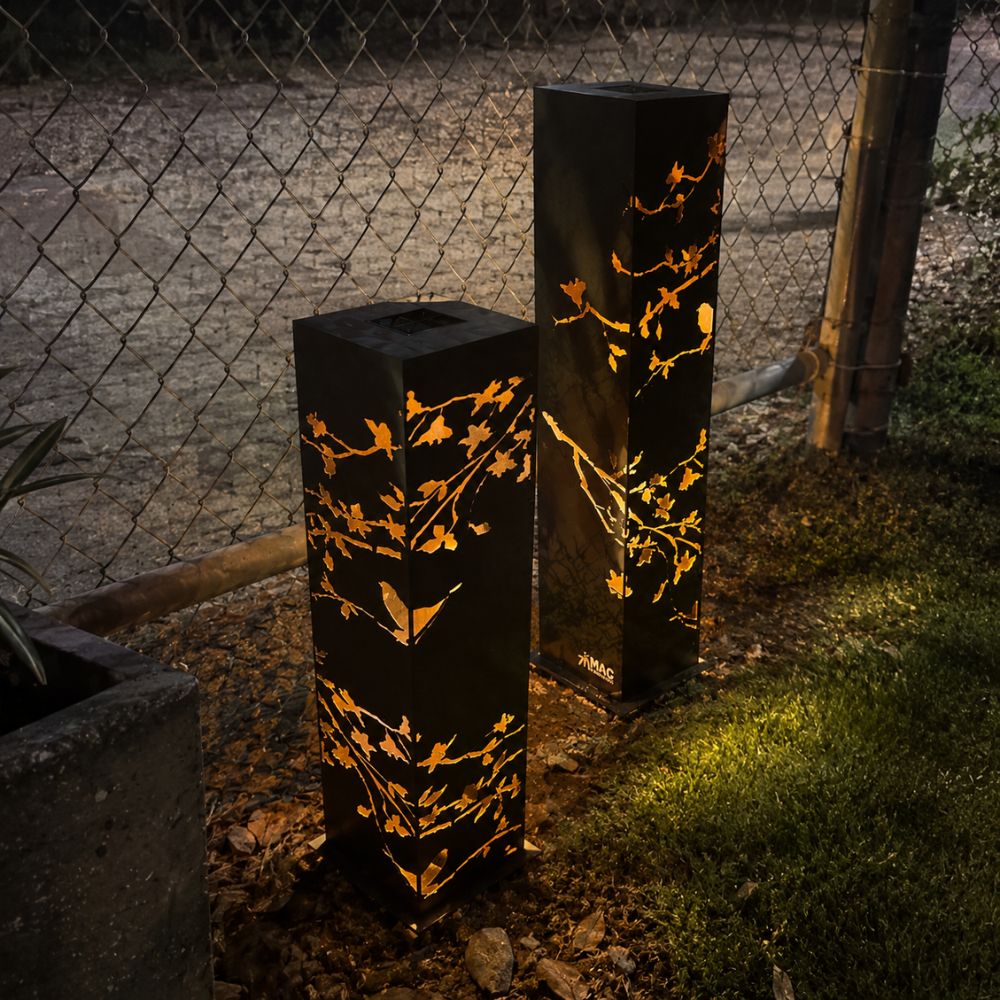 Corten Steel Solari Lamp in a contemporary outdoor garden at night, featuring illuminated rust-finished steel pillar lights with intricate laser-cut leaf patterns. The warm internal lighting casts decorative shadows, creating an elegant ambience for modern landscaping and garden pathways.