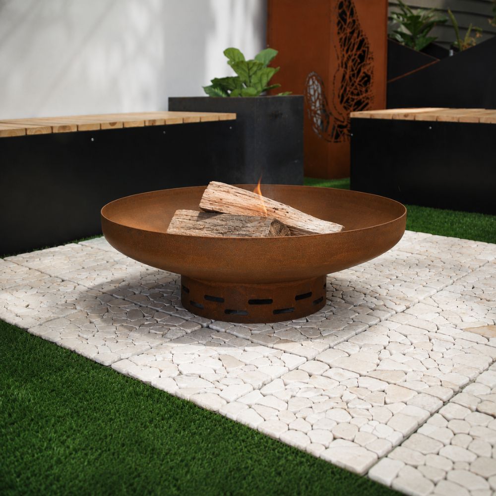 A Corten steel terra fire pit in a modern outdoor courtyard, featuring a wide rust-finished steel bowl with burning firewood, set on a light stone-paved area and surrounded by timber benches, planters, and decorative Corten steel privacy screens.