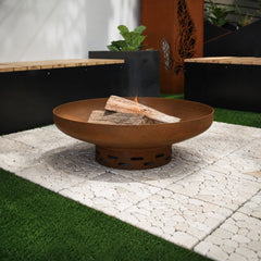 A Corten steel terra fire pit in a modern outdoor courtyard, featuring a wide rust-finished steel bowl with burning firewood, set on a light stone-paved area and surrounded by timber benches, planters, and decorative Corten steel privacy screens.