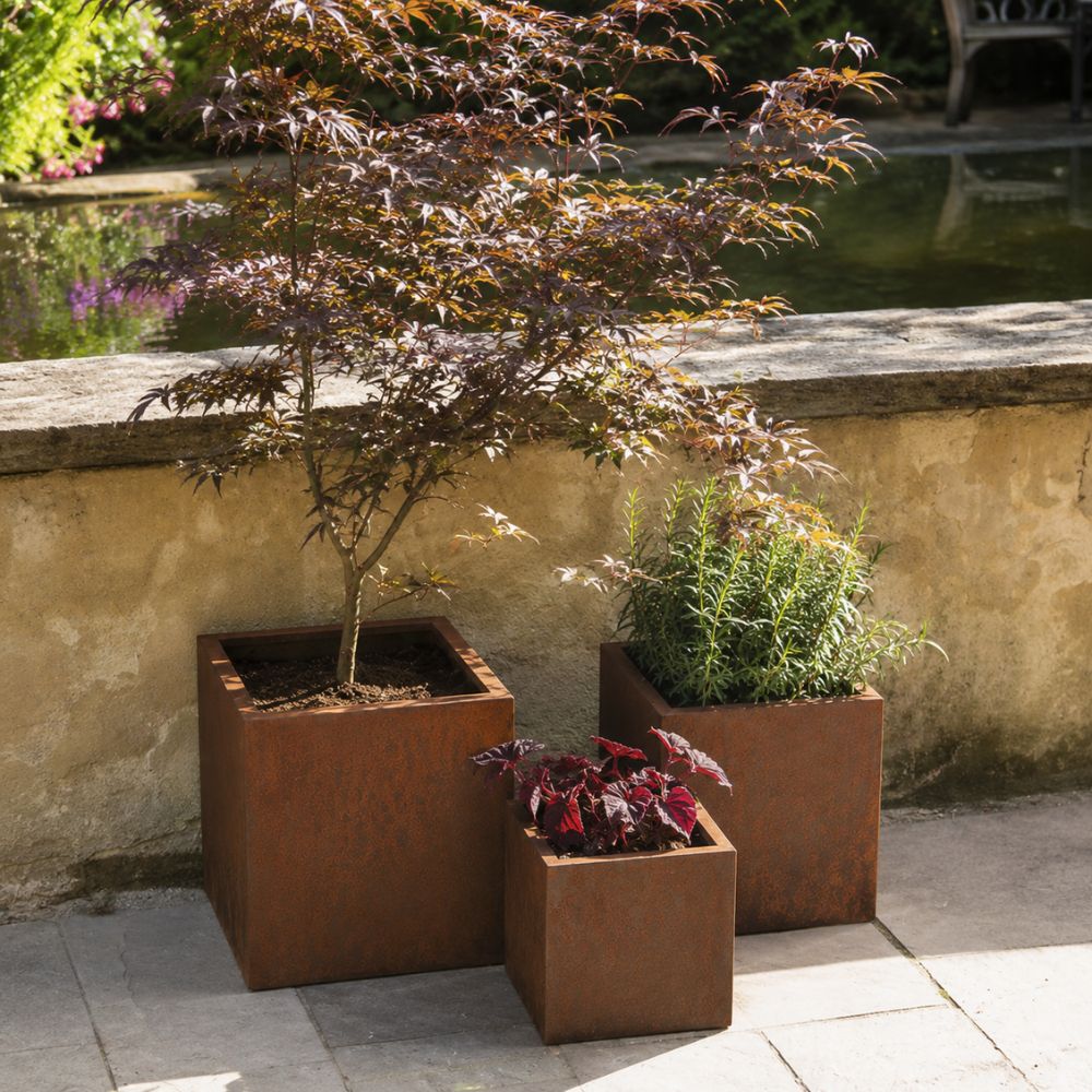 Corten Steel VitaSquare Classic Plant Pot set in a landscaped outdoor garden, featuring rust-finished square steel planters in multiple sizes. Filled with ornamental trees, shrubs, and colourful foliage, the planters add a contemporary architectural touch beside a stone patio and tranquil garden pond.