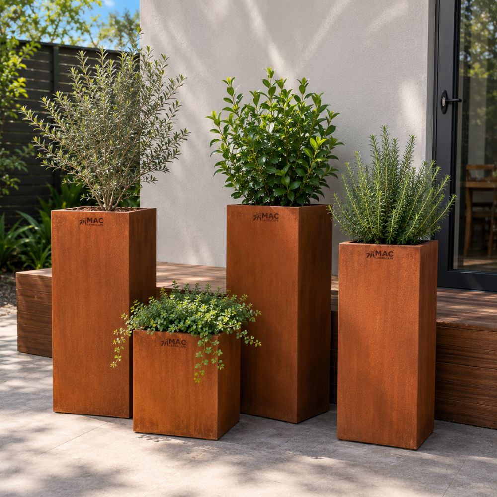 Corten Steel Podium Planter collection featuring tall, rust-finished steel planters in multiple sizes. Filled with ornamental shrubs and herbs, the contemporary rectangular planters create an elegant architectural display on a modern outdoor patio beside a residential entrance.