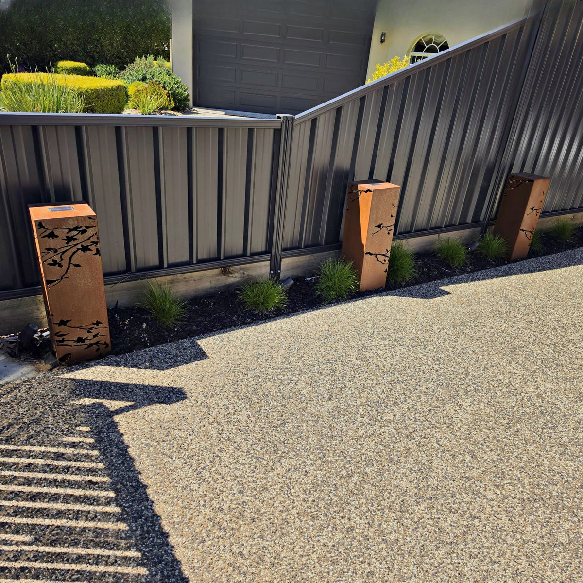 Customer photo: three Corten Steel Solari Lamps with leaf cut-out panels lining a garden bed beside a driveway